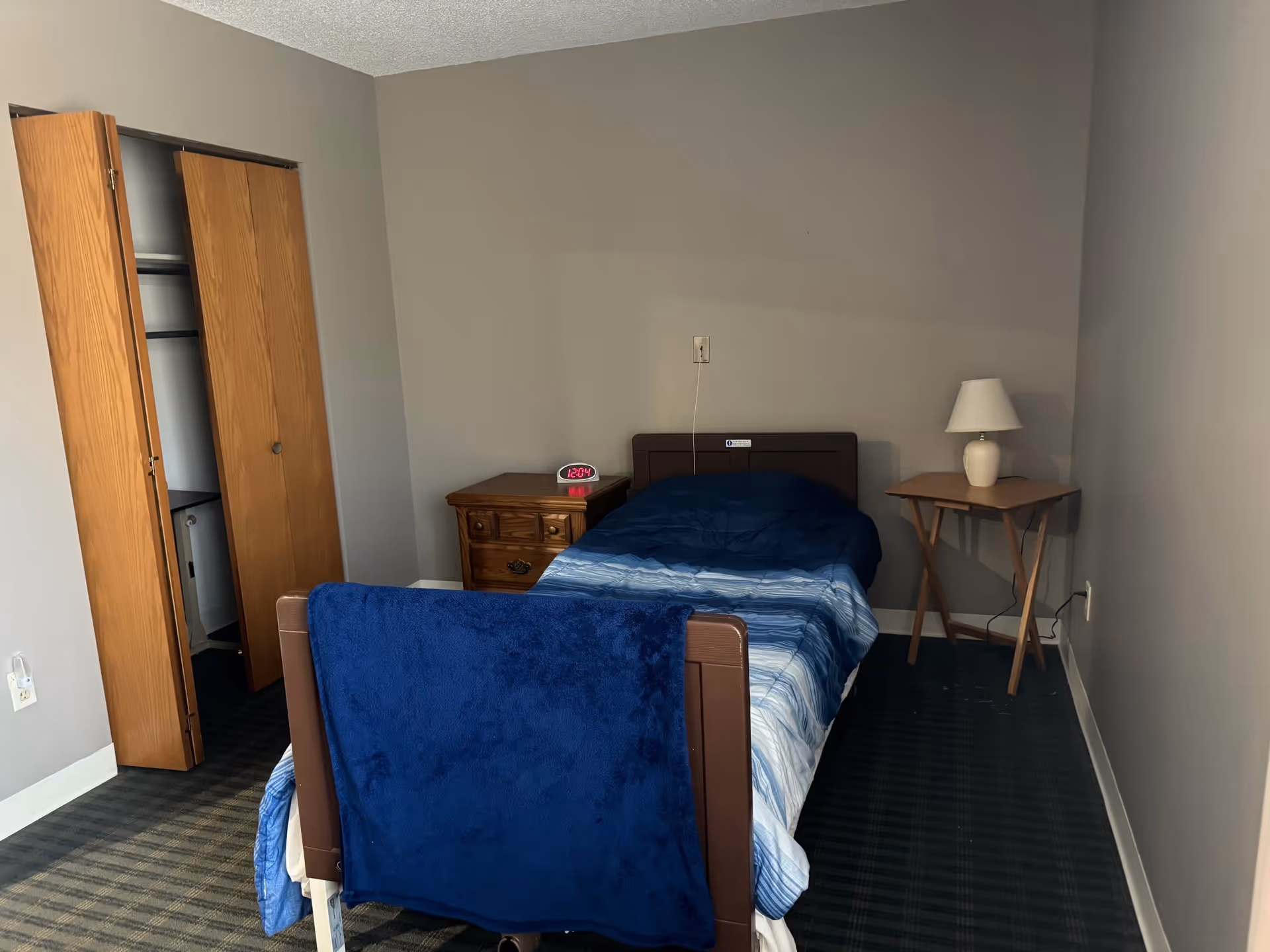 Small bedroom with a single bed covered in blue bedding, a nightstand with a clock, a side table lamp, and an open closet.