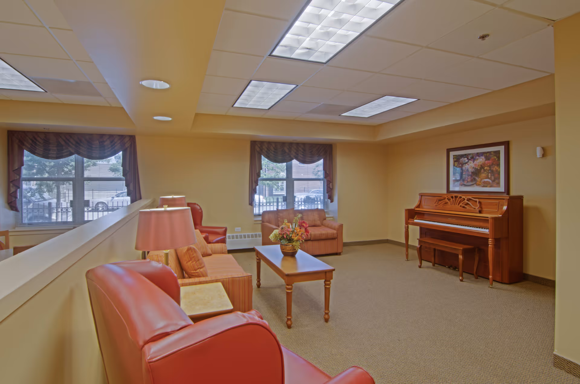 A cozy senior living common area with beige walls and carpet. The room features a wooden piano with a bench, a framed floral painting above it, two windows with valance curtains, a wooden coffee table with a flower arrangement, and several upholstered chairs and sofas in warm tones. Ceiling lights illuminate the space.