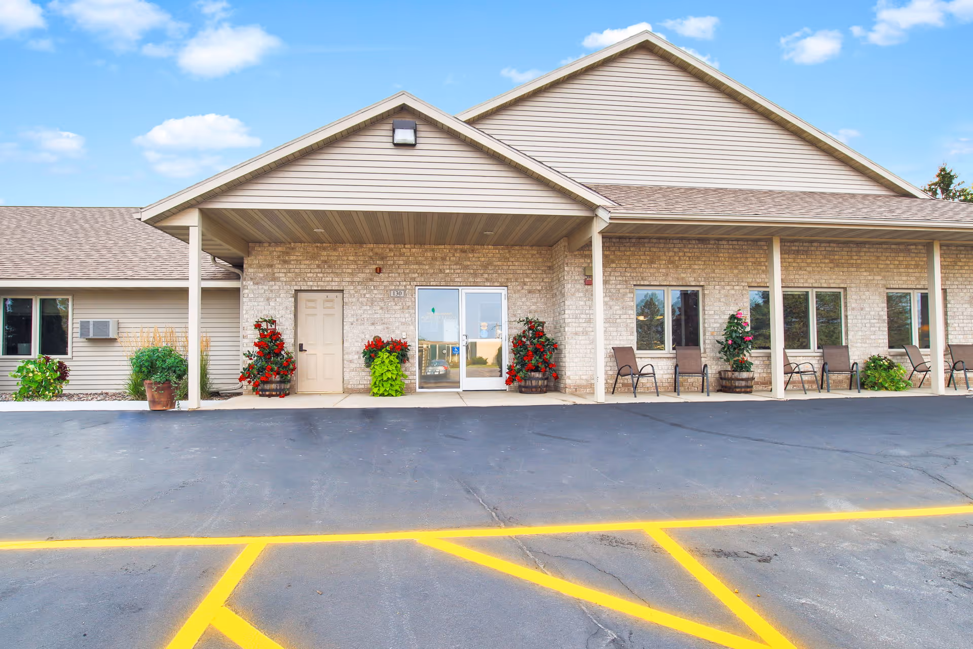 Front entrance of a single-story assisted living building with a covered porch, glass doors, potted plants, and outdoor seating.