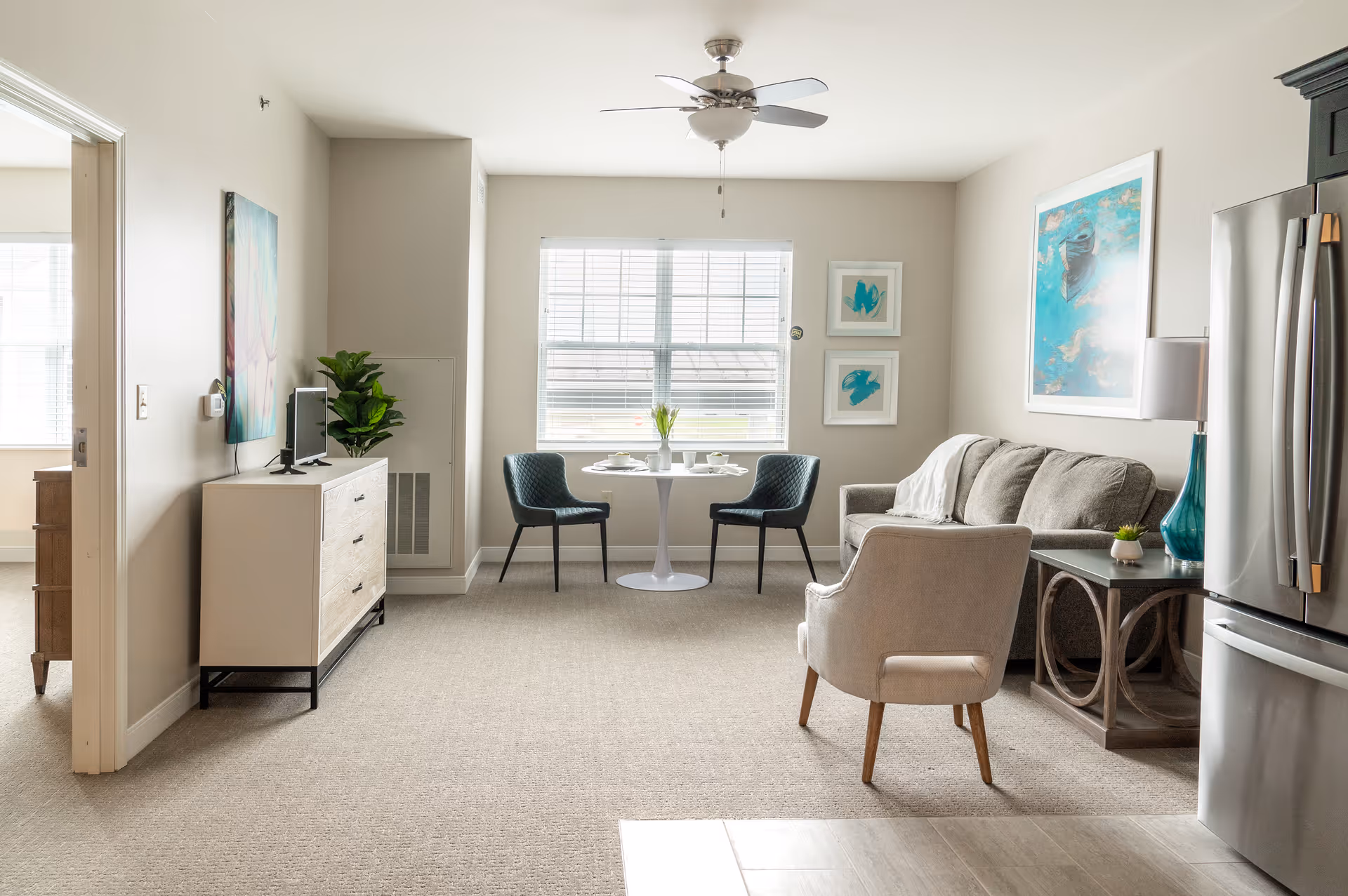 A bright and modern living room area with a beige sofa, a light-colored armchair, a side table with a blue lamp, and a small round dining table with two dark chairs near a window. A ceiling fan is mounted above, and a stainless steel refrigerator is visible on the right side. The room has light-colored walls and carpeted flooring, with artwork hanging on the walls.