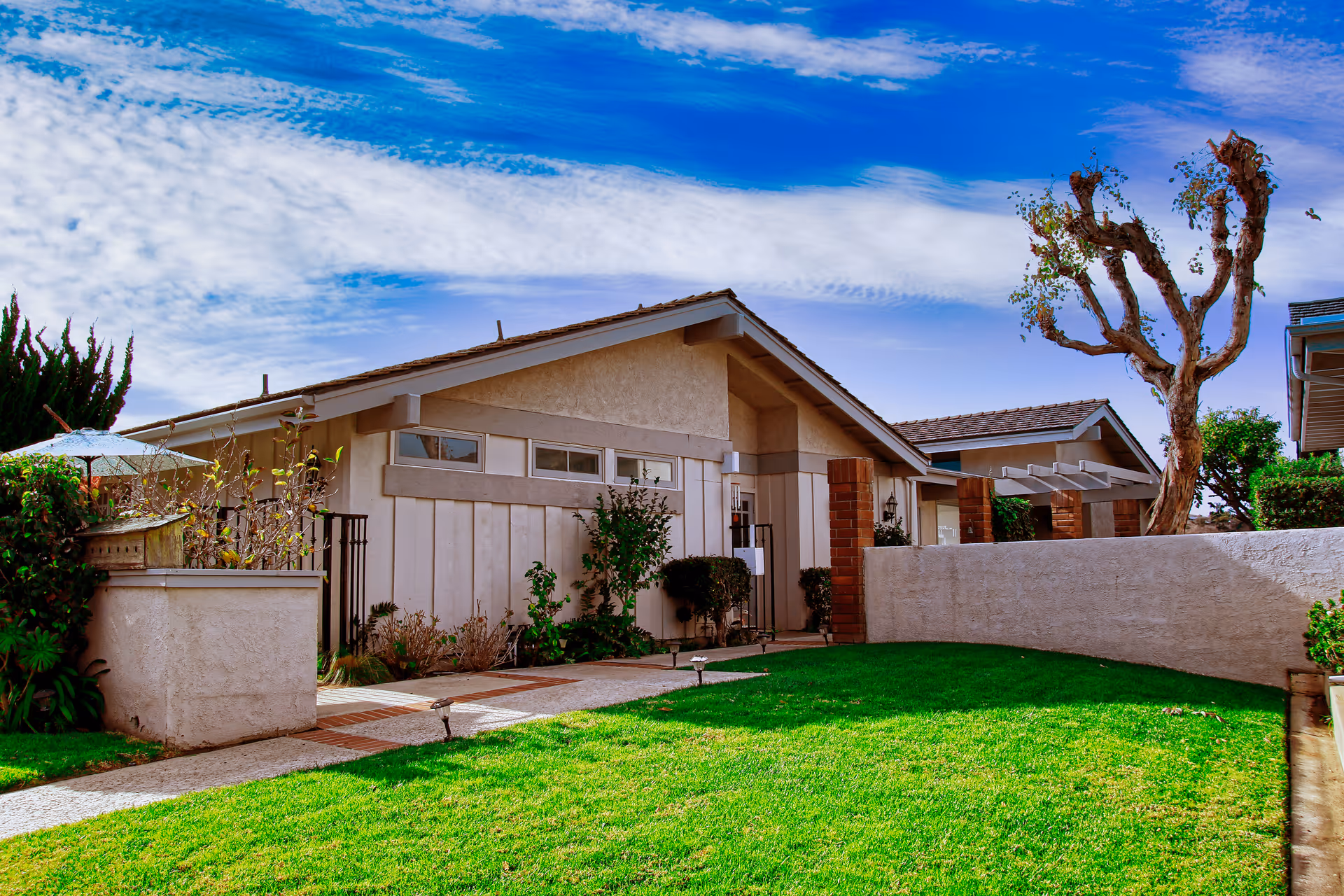 Exterior view of a single-story residential building with beige walls and a sloped roof under a partly cloudy blue sky. The building is surrounded by a well-maintained green lawn, some bushes, and a few trees, including one with pruned branches. A paved walkway leads to the entrance of the building.