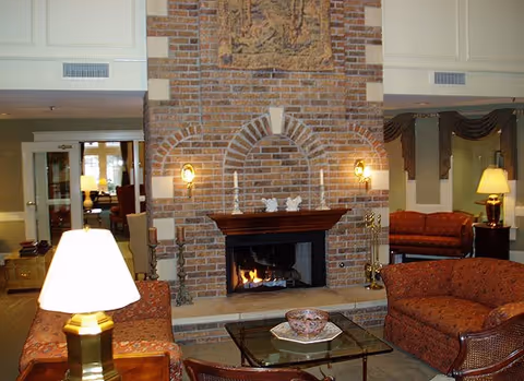 A cozy living room area with a large brick fireplace in the center. The fireplace has a wooden mantle with decorative items and two wall-mounted lamps on either side. In front of the fireplace is a glass coffee table with a decorative bowl. Surrounding the table are upholstered chairs and sofas in warm tones. There are lamps providing soft lighting and a glimpse of other seating areas in the background.