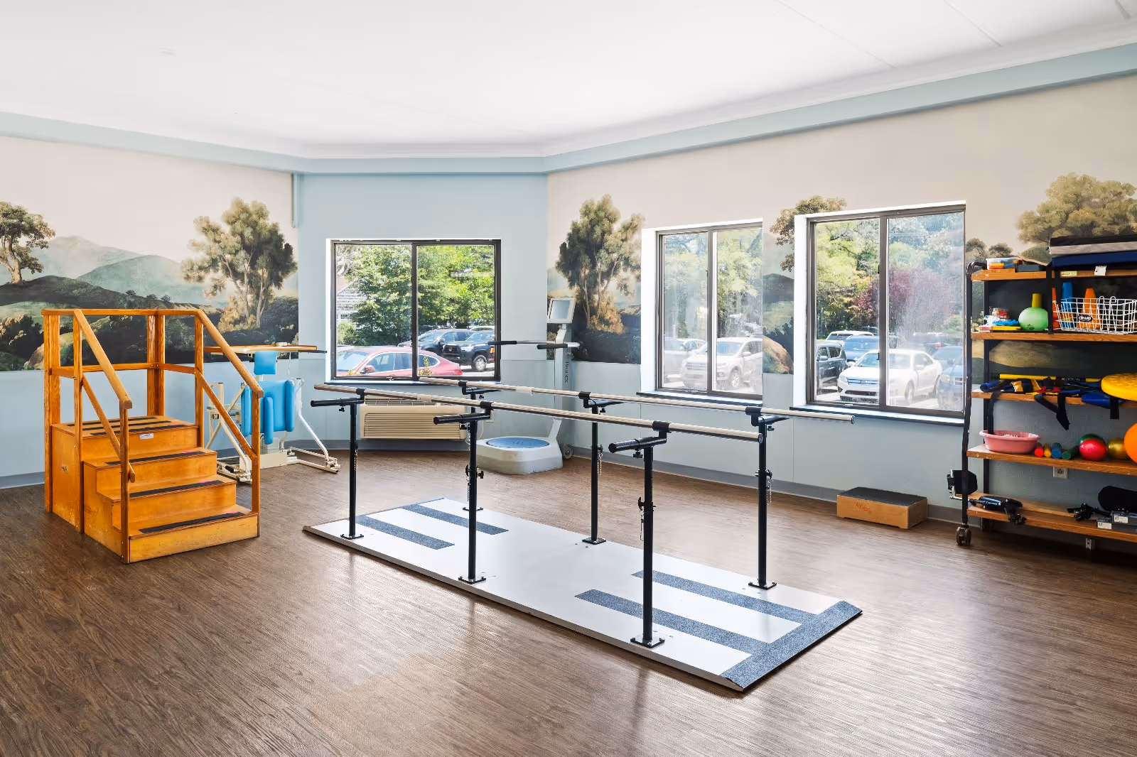 A spacious rehabilitation room with wooden stairs for physical therapy, parallel bars for walking exercises, and various therapy equipment on shelves. The room has large windows letting in natural light and a mural of trees and hills on the walls.