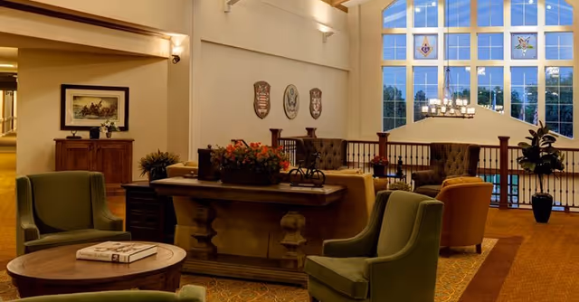Spacious senior living lounge with upholstered armchairs and tables arranged around a central console, large arched windows and a chandelier.