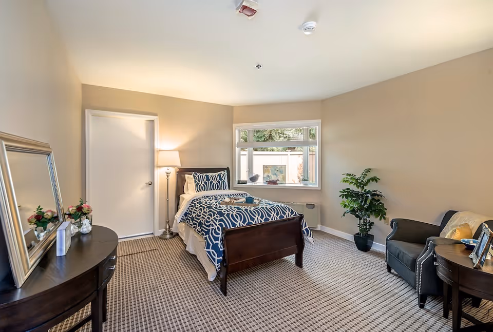 A cozy bedroom with a single bed featuring blue and white patterned bedding, a bedside lamp, a window with a view of greenery outside, a dark wooden dresser with a mirror and flowers, a comfortable armchair, a small round table with picture frames, and a potted plant in the corner.