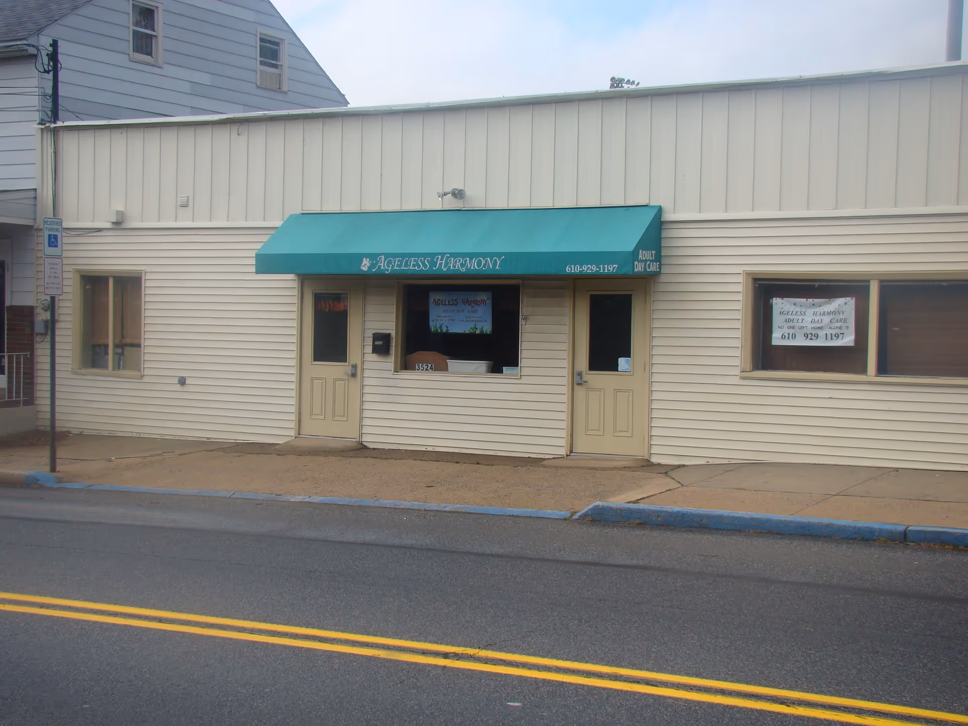 Exterior front view of Ageless Harmony adult day care facility with beige siding, two beige doors, a green awning with the facility name and phone number, and windows displaying signs.