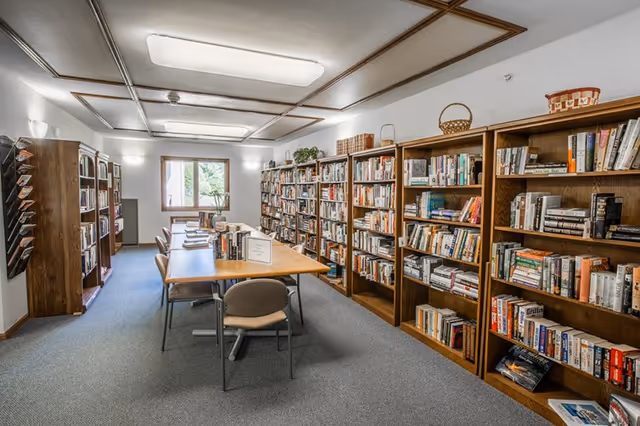 Community library room with wooden bookshelves lining the walls and a central table with chairs under ceiling lights.