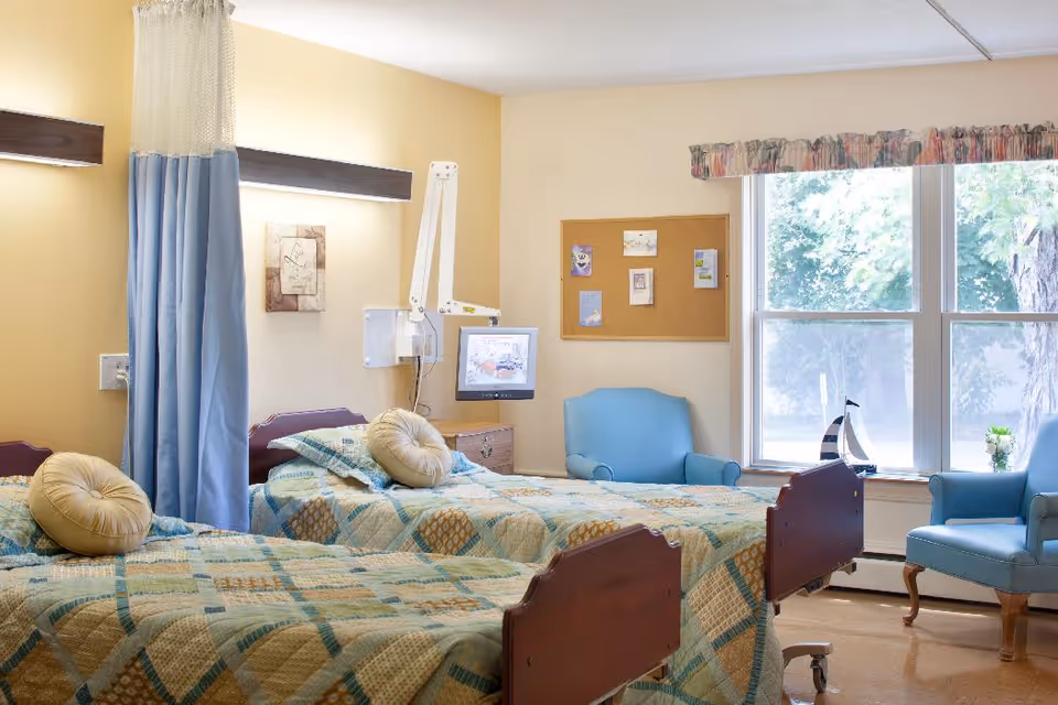 A bright room with two hospital-style beds separated by a blue privacy curtain. Each bed has a patterned quilt and a round beige pillow. There is a blue armchair near a large window with a view of trees outside. A cork bulletin board with pinned notes is on the wall, and a small TV is mounted near one bed.