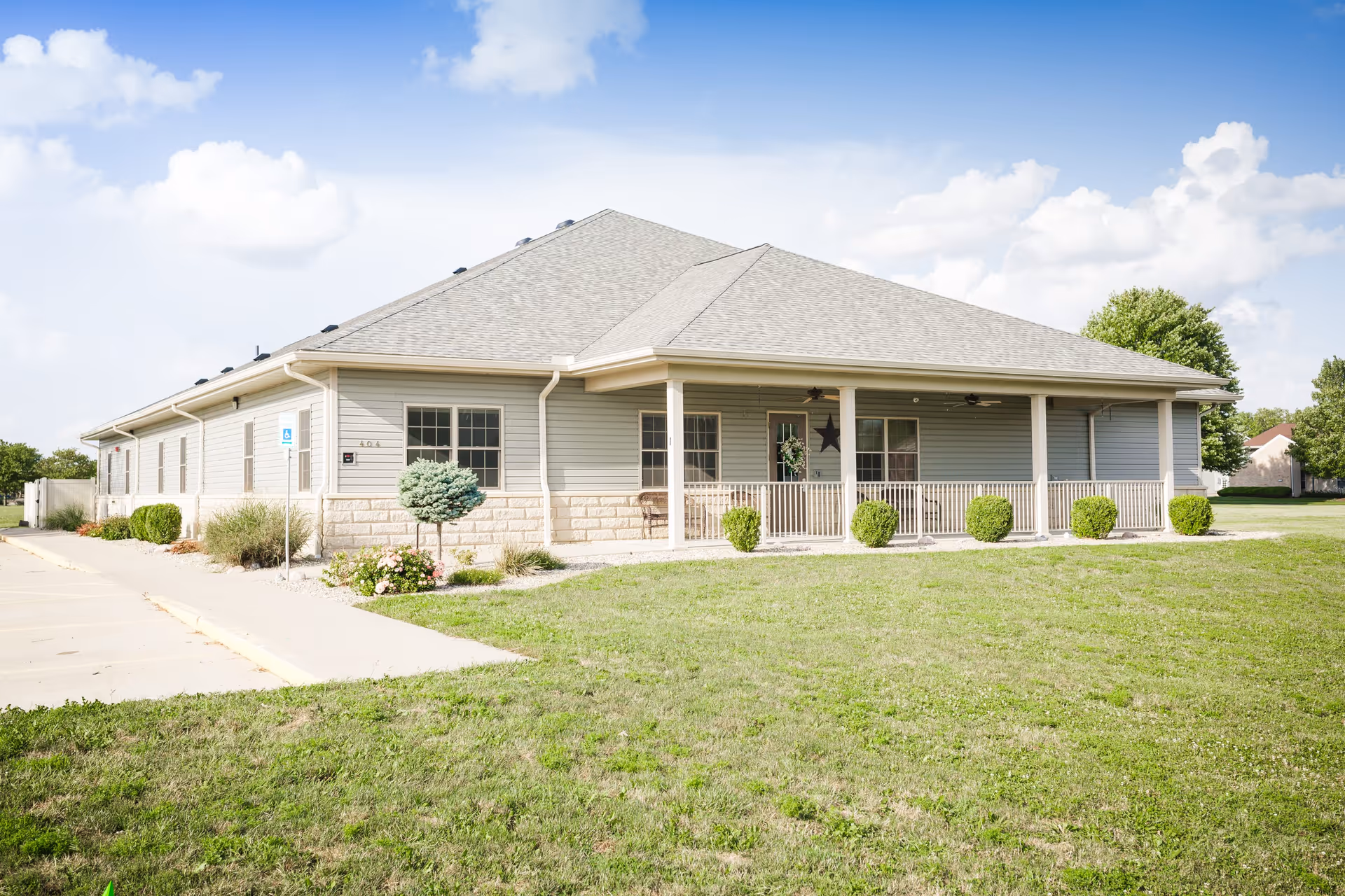 Single-story building with light gray siding and a covered porch with white railings, surrounded by a grassy lawn and small shrubs under a partly cloudy sky.