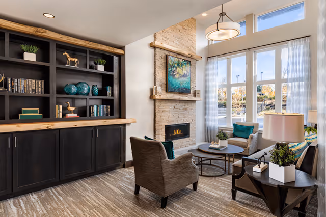 A cozy living room area with a stone fireplace, a painting above the fireplace, large windows with sheer curtains letting in natural light, two armchairs, a sofa with cushions, a round coffee table, and a built-in black bookshelf with decorative items and books.