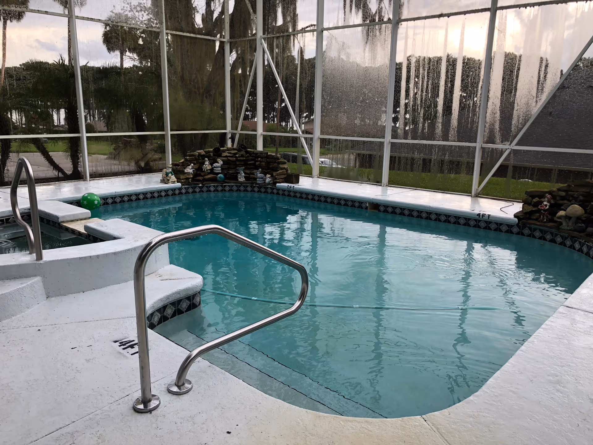 Indoor swimming pool with clear blue water, surrounded by a white deck and metal handrails. The pool area is enclosed with large glass panels, showing trees and greenery outside. There are small decorative statues on the far side of the pool.