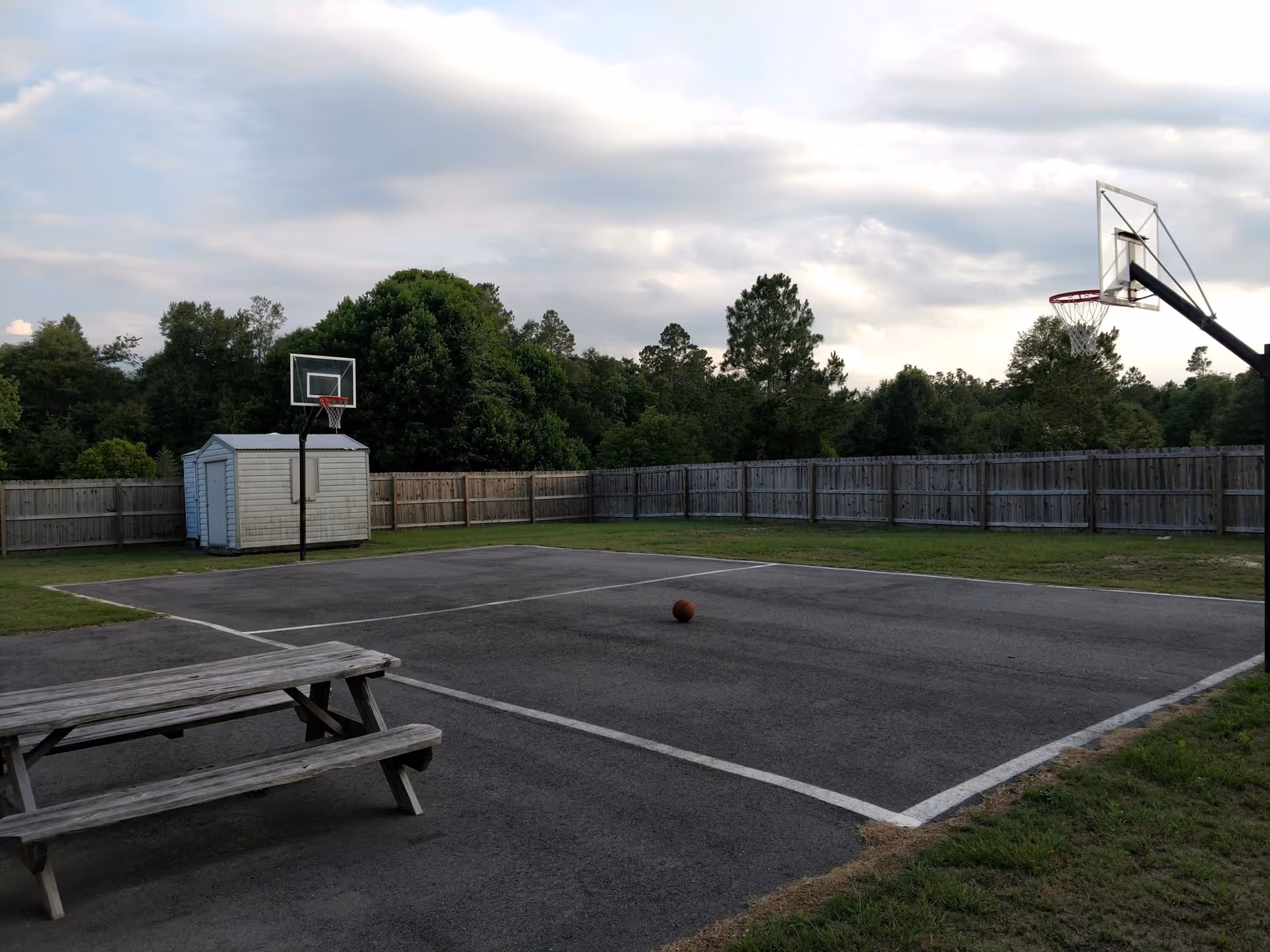 Outdoor fenced basketball court with two hoops, a picnic bench, and a basketball on the pavement.