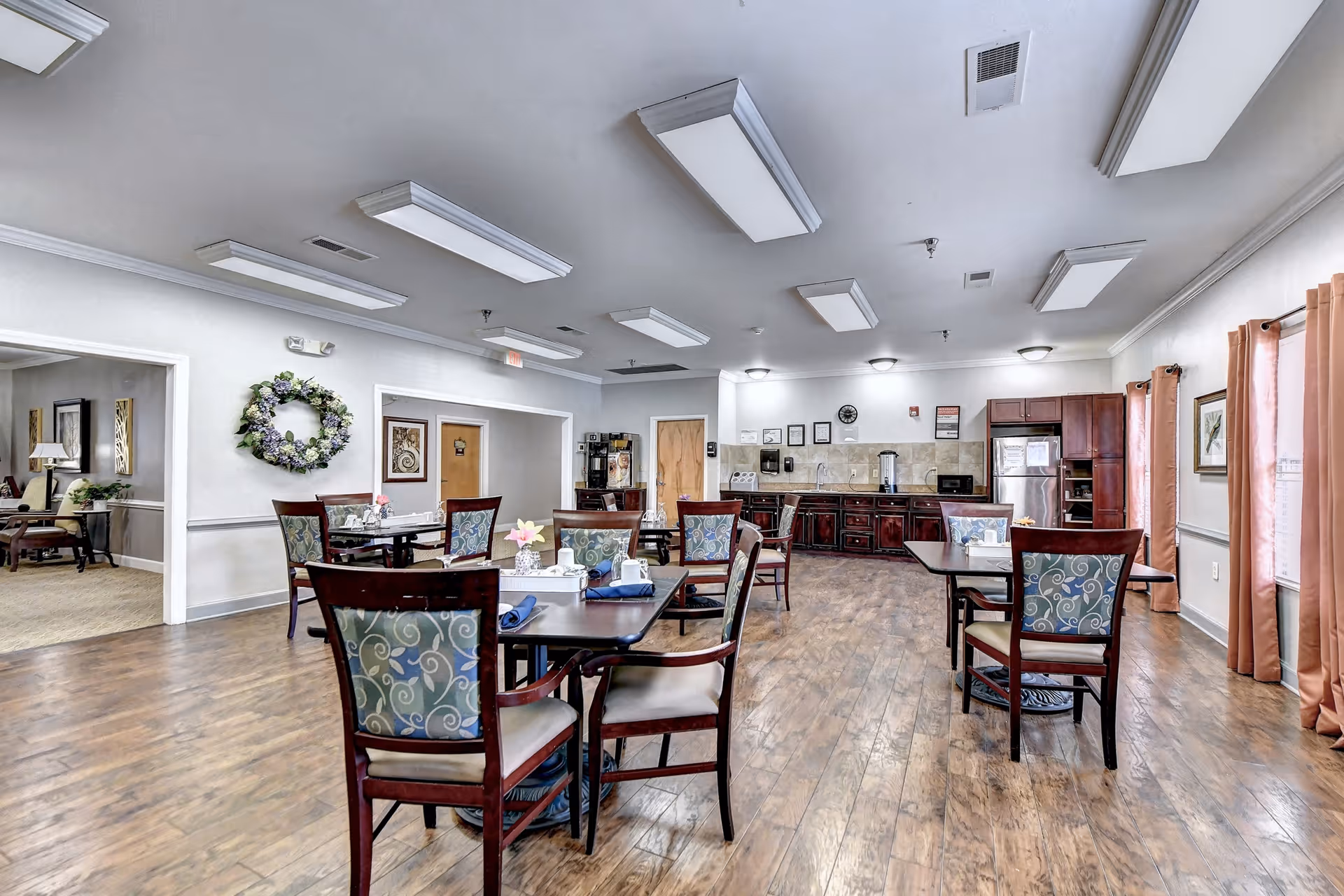 Spacious dining room in a senior living facility with multiple wooden tables and upholstered chairs, wood flooring, and a kitchenette along the back wall.