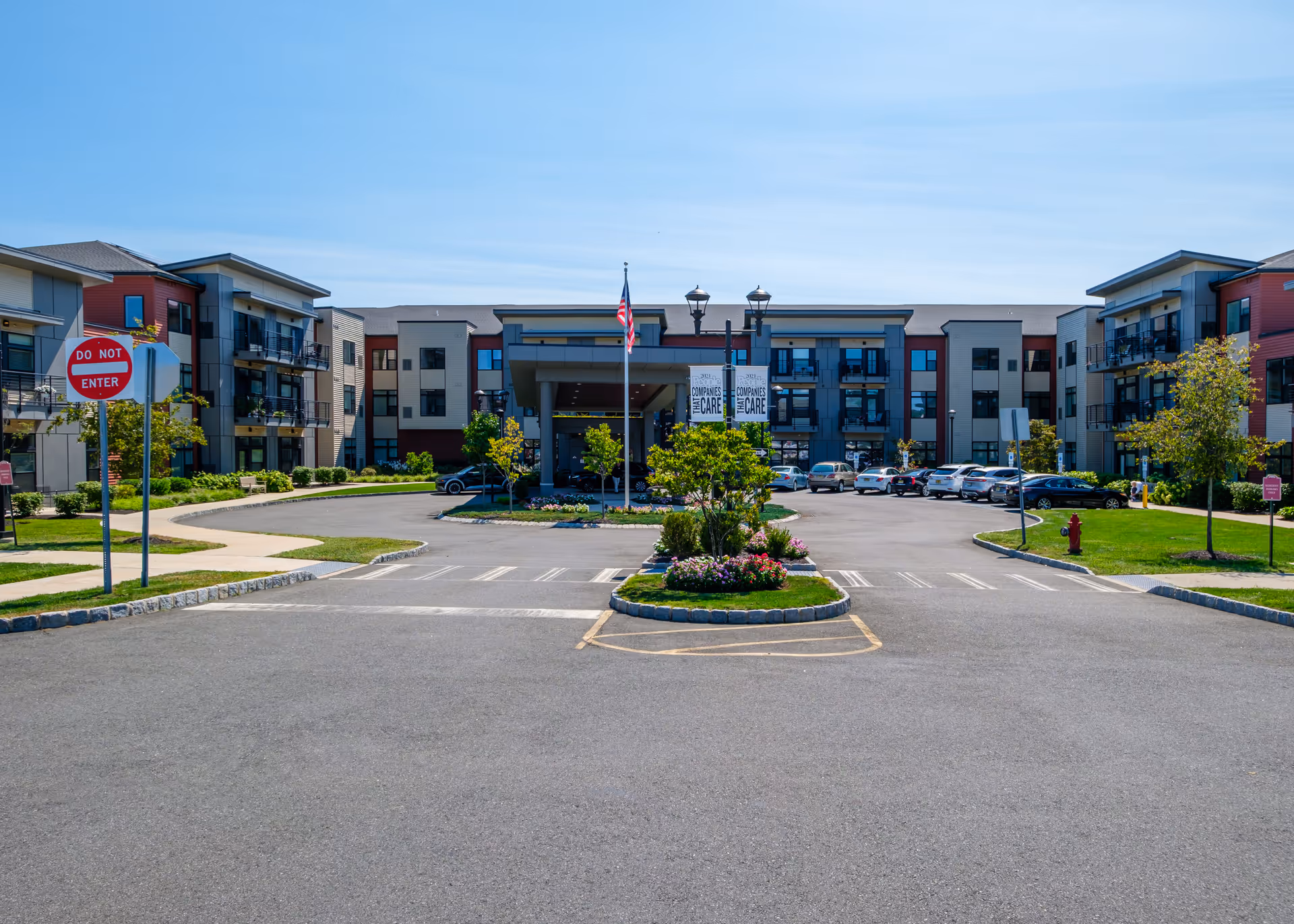 Front exterior view of a senior living facility with a circular driveway, landscaped greenery, an American flag, and multiple parked cars under a clear blue sky.