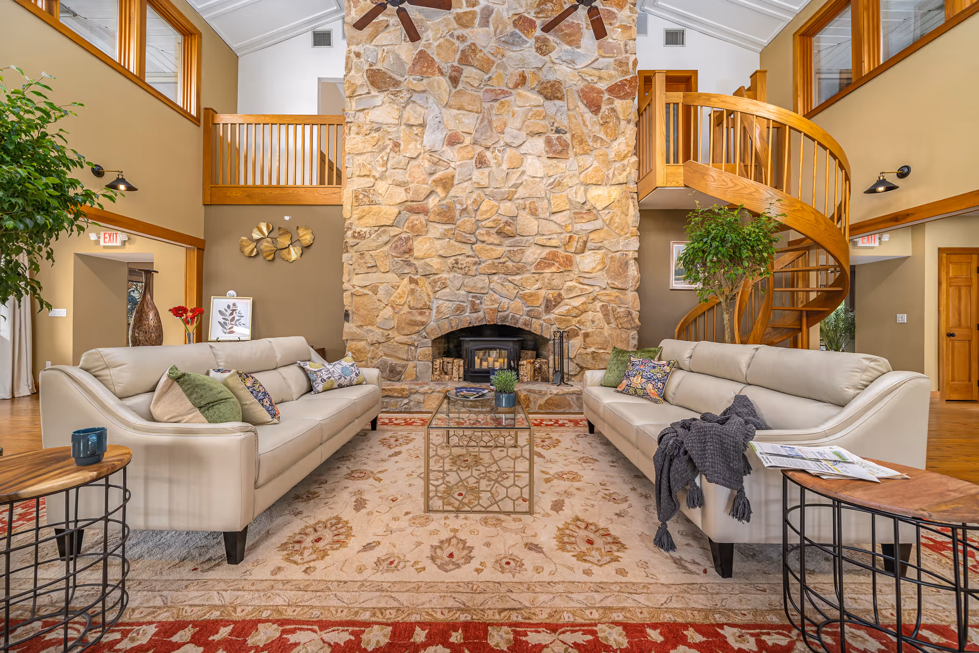 A spacious living room with two beige leather sofas facing each other, a glass coffee table in the center, and a large stone fireplace as the focal point. The room features a high ceiling with wooden beams, large windows, a spiral wooden staircase on the right, and decorative plants and artwork on the walls.