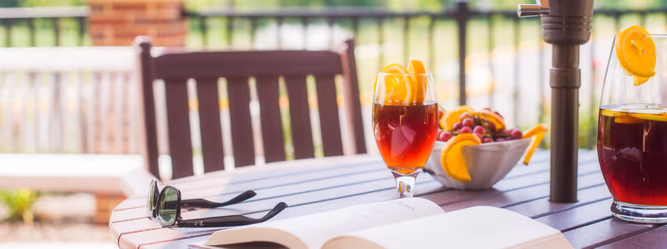 Outdoor patio table with an open book, sunglasses, a glass of iced tea with orange slices, a pitcher and a bowl of fruit, with wooden chairs in the background.