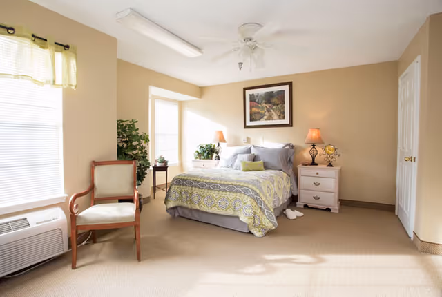 Sunlit bedroom with a double bed dressed in patterned bedding, nightstands with lamps, an armchair, ceiling fan, and windows.