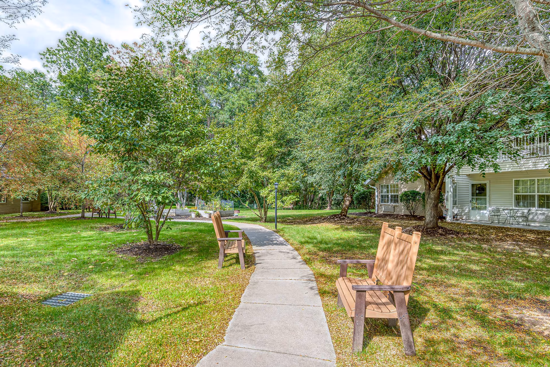 A peaceful outdoor garden area at Solstice Senior Living at Fairport featuring a curved concrete walkway flanked by wooden benches, surrounded by lush green grass, trees, and shrubs. A building with white siding and windows is visible on the right side under the shade of trees.