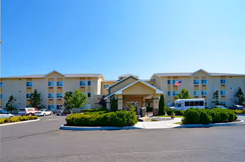 Exterior view of a three-story senior living facility building with a central covered entrance, surrounded by landscaped bushes and trees. Several cars and a white shuttle van are parked in front of the building under a clear blue sky.