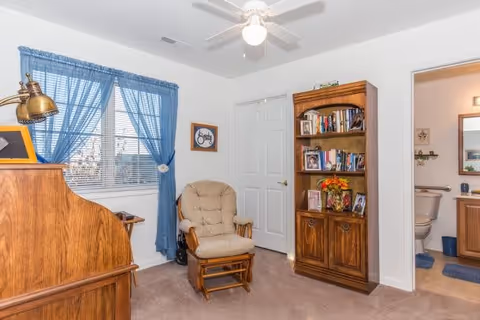 A cozy room with a wooden rocking chair with beige cushions, a wooden bookshelf filled with books and framed photos, a wooden desk with a brass lamp, a window with blue curtains, and a ceiling fan. An open door reveals a bathroom with a toilet and a mirror.