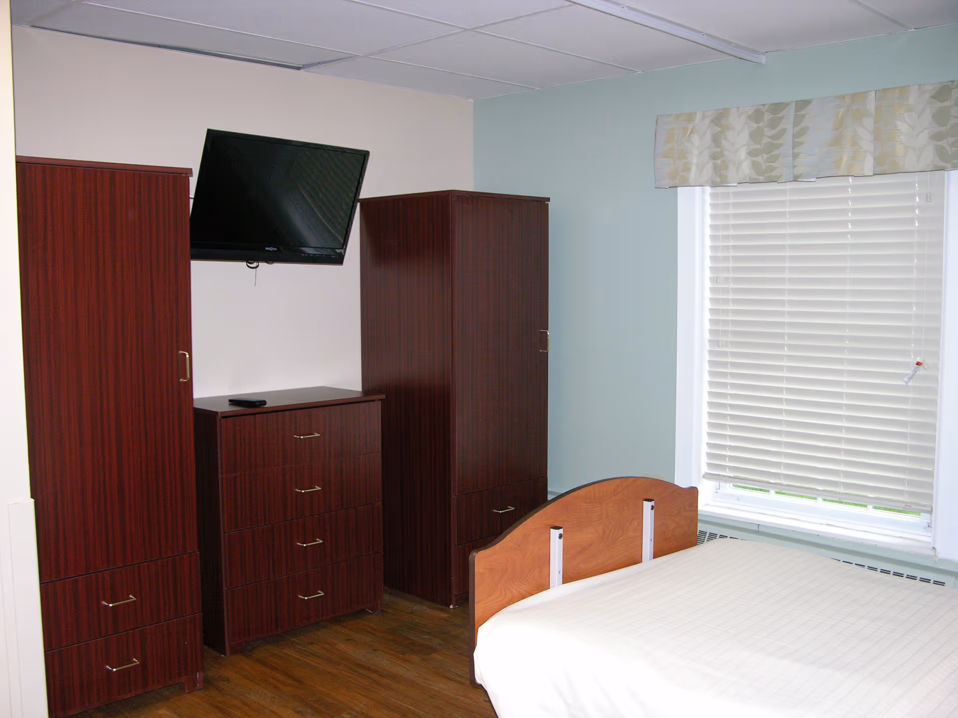 A simple bedroom with a wooden bed frame and a white mattress, two tall wooden wardrobes, a wooden chest of drawers with a remote control on top, a wall-mounted flat screen TV, and a window with closed blinds and a patterned valance.