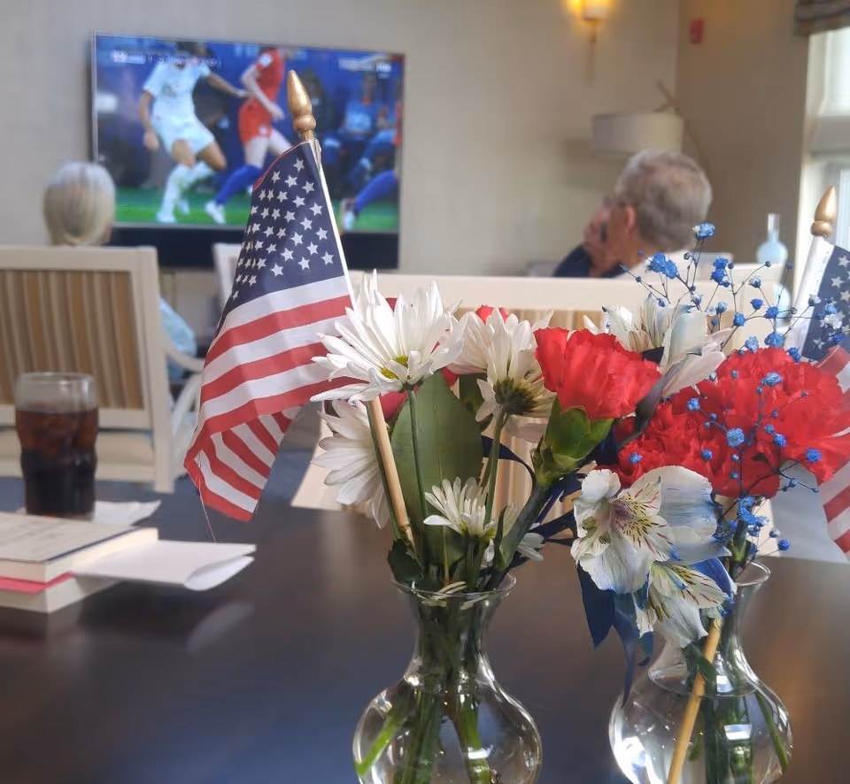 Two small glass vases with red, white, and blue flowers and miniature American flags on a table in a common area. In the background, two elderly people are seated facing a television showing a soccer game.