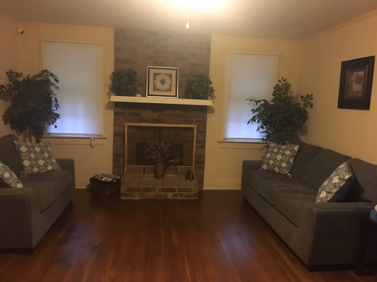 Cozy living room with a brick fireplace centered between two windows, flanked by two gray sofas and potted plants on hardwood floors.