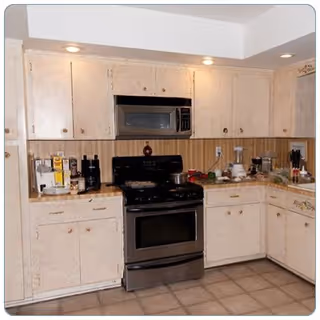 A kitchen with beige cabinets and a black stove with an overhead microwave. The countertop has various kitchen appliances and items including a blender, coffee maker, and some vegetables. The floor is tiled and there are recessed lights in the ceiling.
