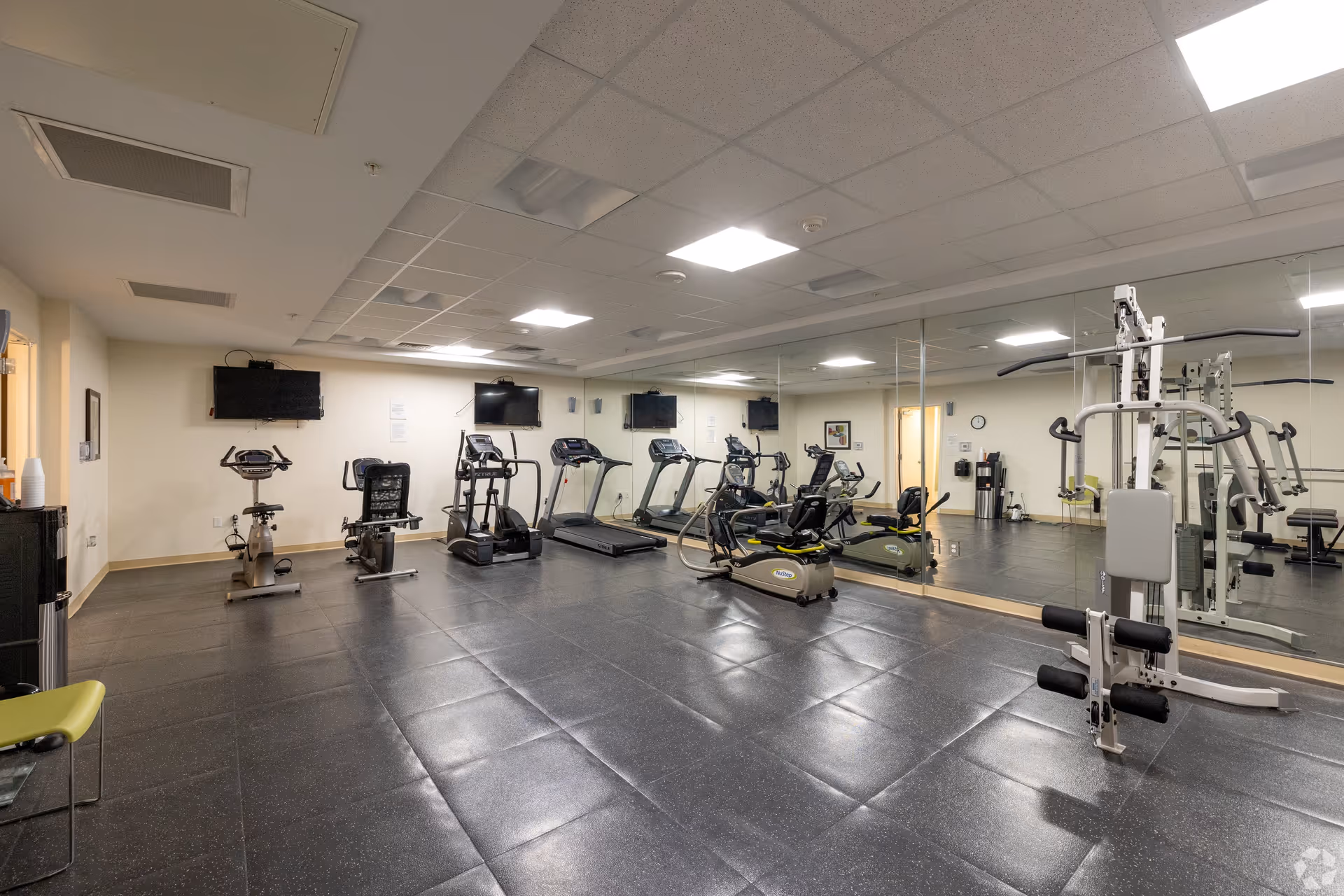 A fitness room with exercise equipment including stationary bikes, treadmills, an elliptical machine, and a multi-function weight machine. The room has large mirrors on one wall, a tiled floor, and bright overhead lighting.
