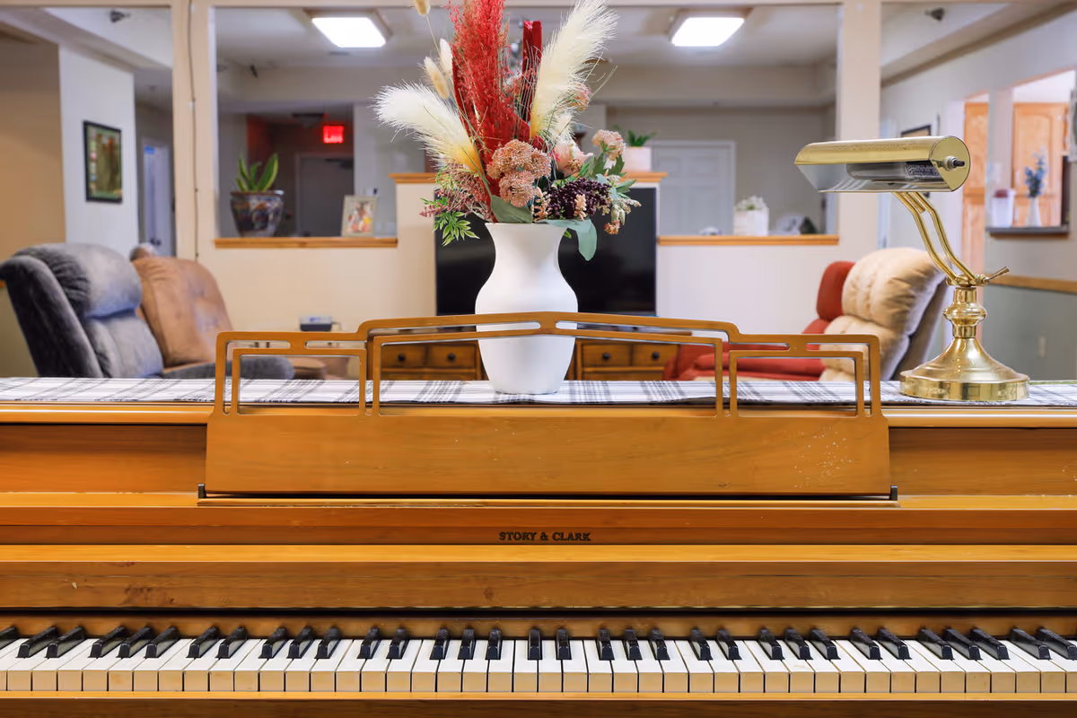 Close-up view of a wooden Story & Clark piano with a white vase holding a floral arrangement on top. Behind the piano, there are several cushioned chairs in a common area with soft lighting and neutral-colored walls.