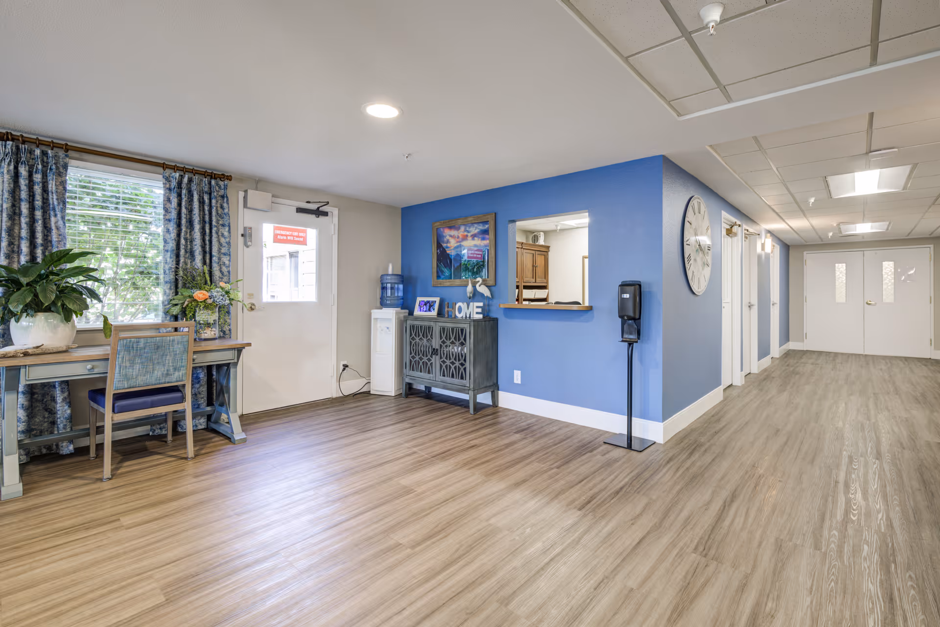 A clean and bright interior hallway area with wood-style flooring and blue accent walls. There is a small table with a chair and plants near a window with blue patterned curtains. A white door with a red emergency exit sign is visible. A water cooler and a decorative cabinet with a framed picture and the word 'HOME' sit against the blue wall. A large clock is mounted on the wall near a hallway with multiple doors and double white doors at the end.