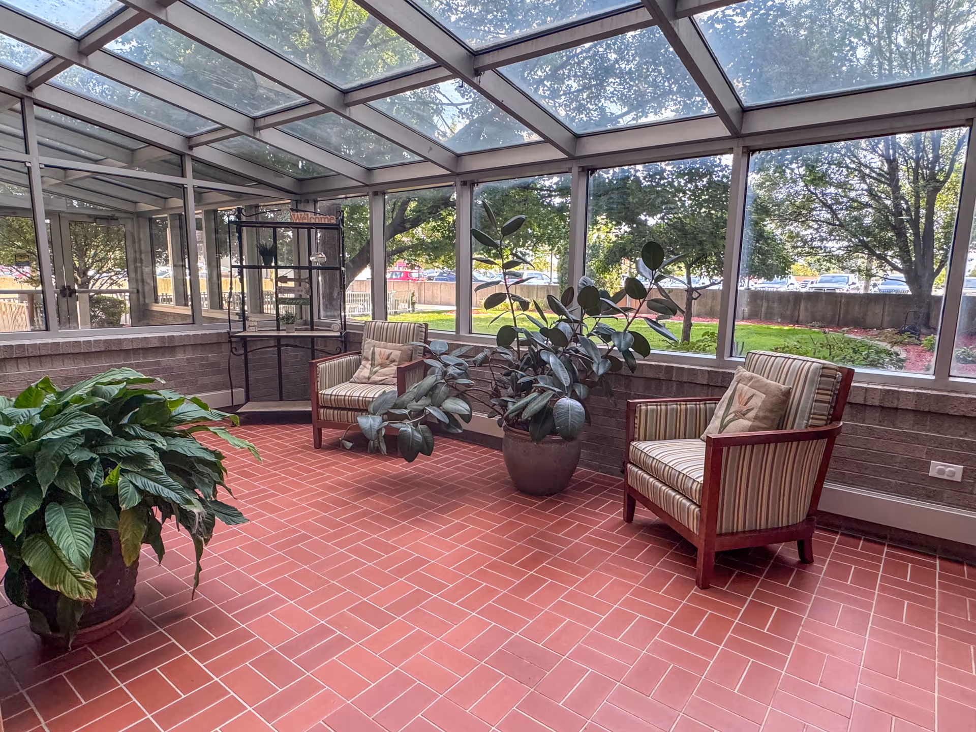 A sunroom with large windows and a glass ceiling, featuring two striped armchairs with cushions, two large potted plants, and a metal shelving unit with a welcome sign. Outside, trees and a grassy area are visible.