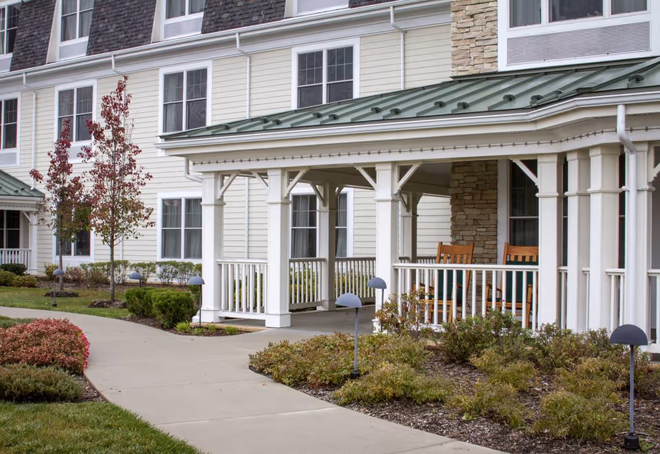 Exterior view of a senior living facility with a covered porch featuring white columns and railings. There are two wooden rocking chairs on the porch, surrounded by landscaped bushes and small trees with red and green foliage. The building has light-colored siding and multiple windows.