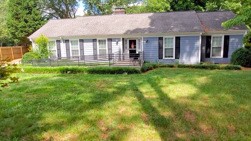Single-story blue house with white trim and black shutters, featuring a front door with a small porch and a wheelchair ramp. The house is surrounded by a green lawn and some bushes, with trees in the background.