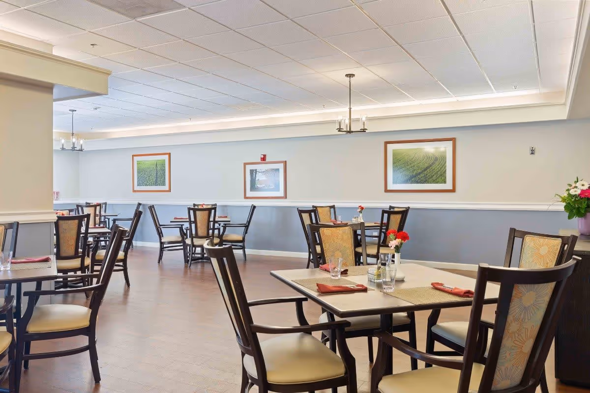A dining room in a senior living facility with multiple tables and chairs arranged neatly. Each table has placemats, glasses, napkins, and small flower vases. The walls are decorated with framed landscape pictures, and the room has a clean, bright atmosphere with overhead lighting fixtures.