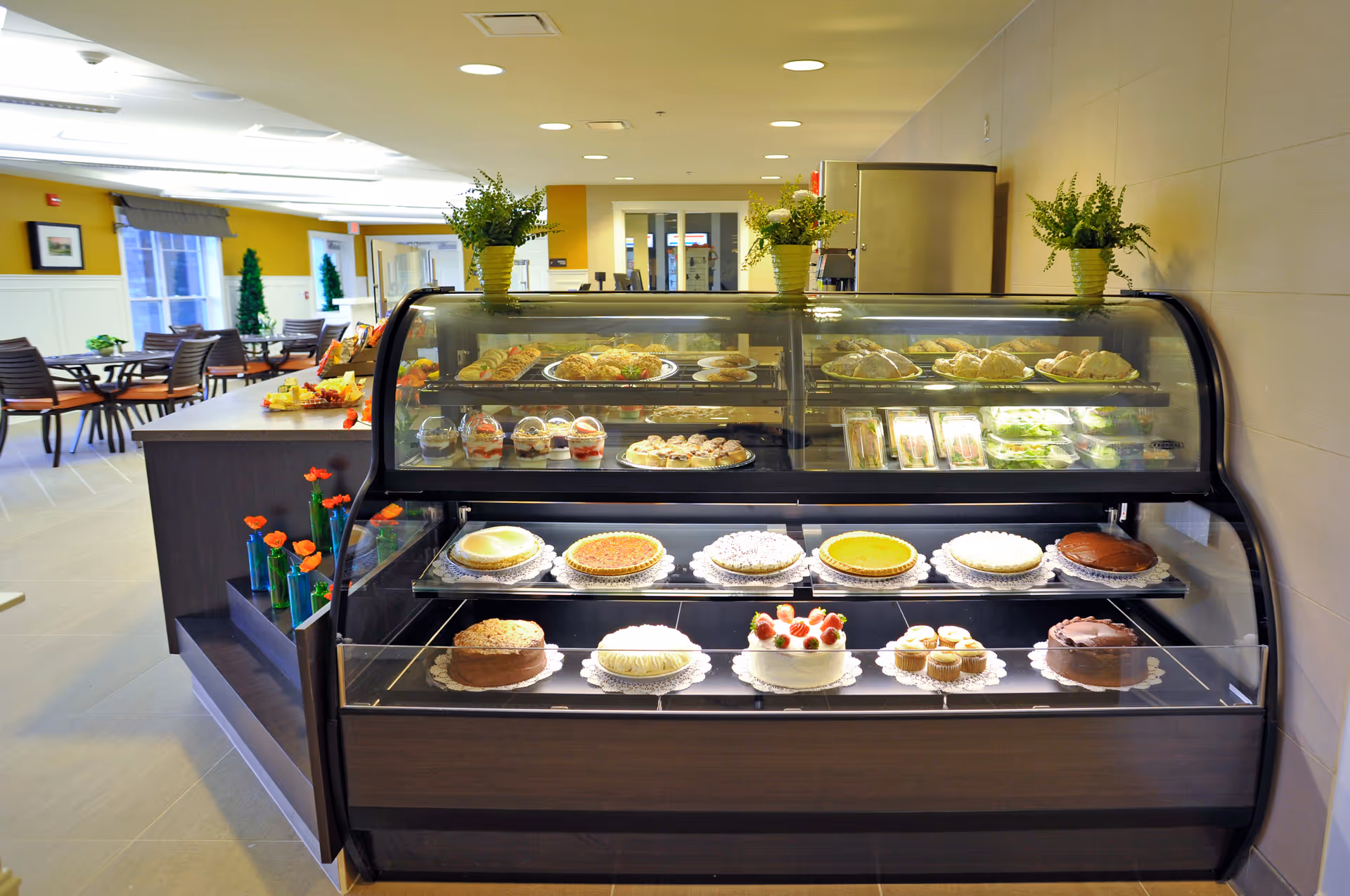 Glass display case filled with cakes and pastries in a bright dining area with tables and chairs.