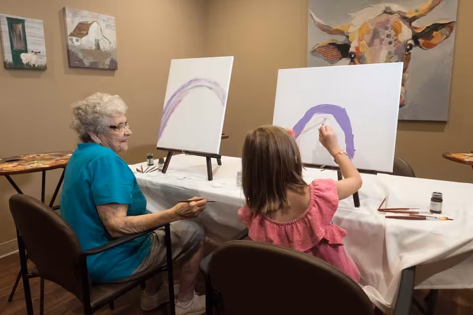 An elderly woman and a young girl seated at a table painting on canvases in a room decorated with animal paintings on the walls.