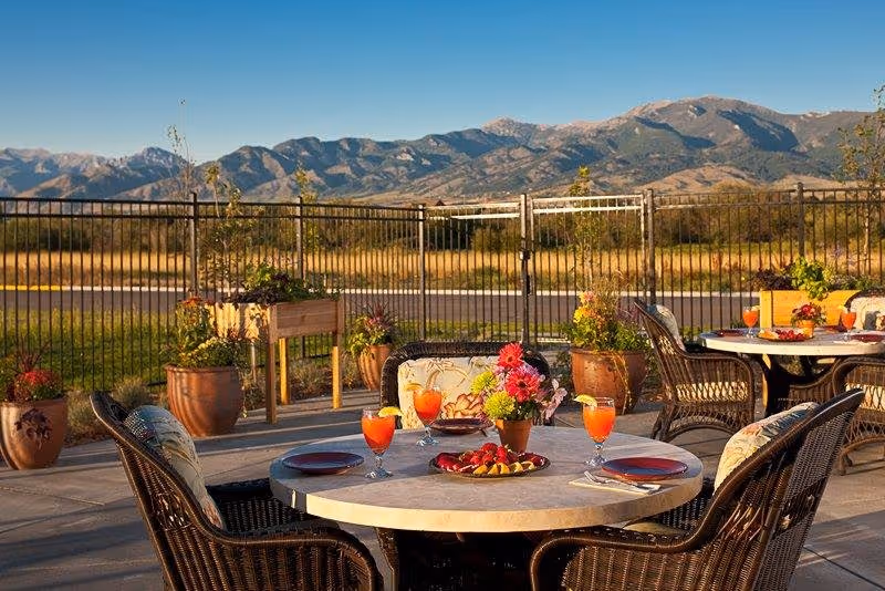 Outdoor patio area with round tables set for dining, featuring wicker chairs with cushions, plates, glasses of orange beverage, and a centerpiece of flowers. The patio is enclosed by a metal fence, with potted plants and a raised garden bed. In the background, there is a scenic view of mountains under a clear blue sky.