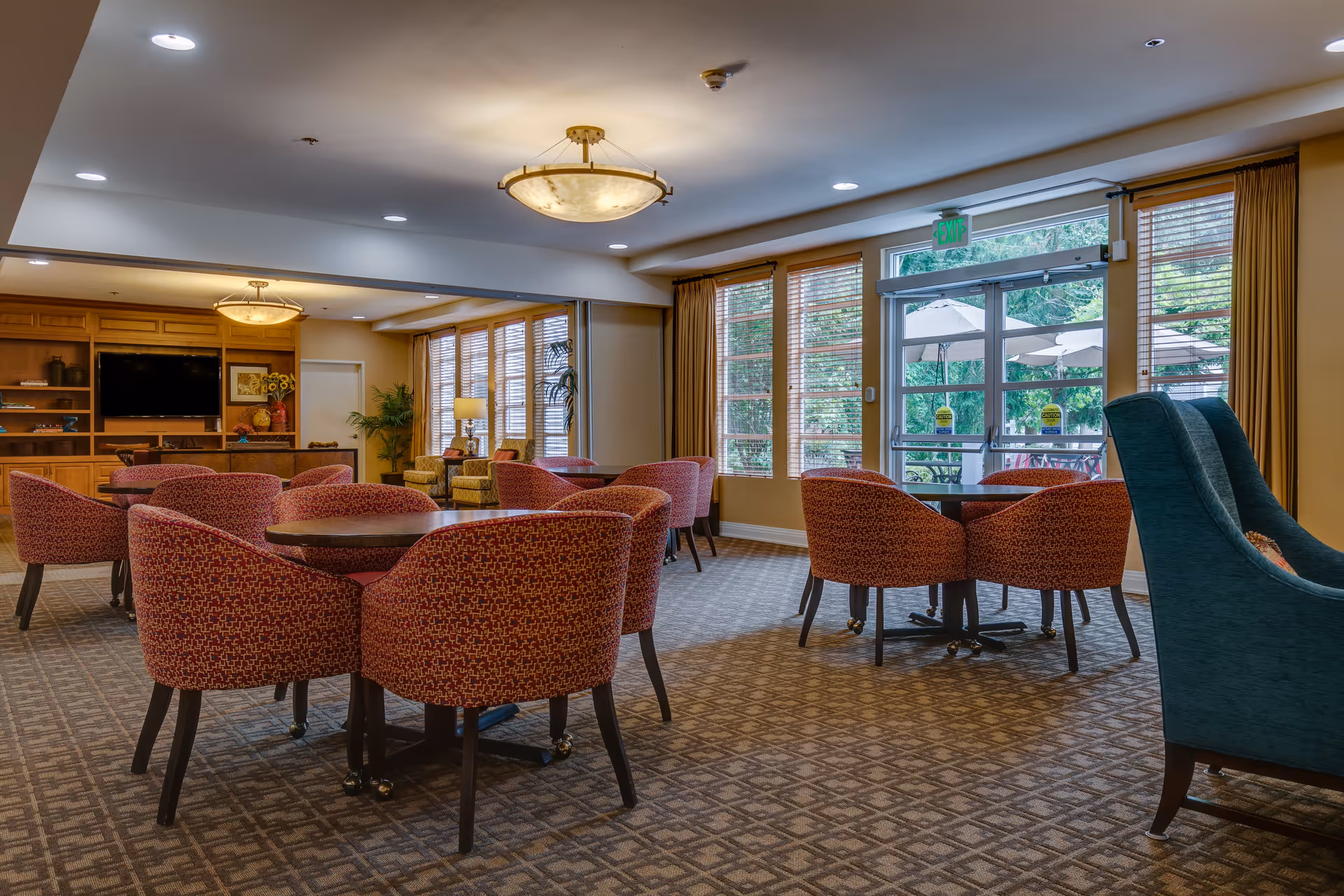 A spacious common area with multiple round tables surrounded by red patterned chairs. The room has large windows with wooden blinds and a glass door leading outside to a patio with umbrellas. There is a built-in wooden entertainment center with a TV and decorative items, and comfortable seating including a blue armchair and two yellow armchairs near the windows. The ceiling has recessed lighting and a central decorative light fixture.