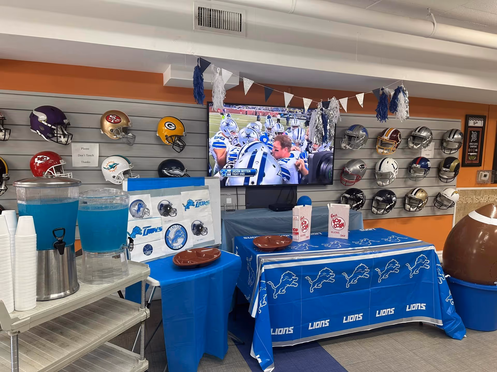 A room decorated for a football event with a Detroit Lions theme. There are multiple football helmets mounted on the wall, a TV showing a football game, tables covered with Detroit Lions tablecloths, popcorn containers, and blue drinks in dispensers with cups stacked nearby. The walls are painted orange and there are blue and white pennant banners hanging above.