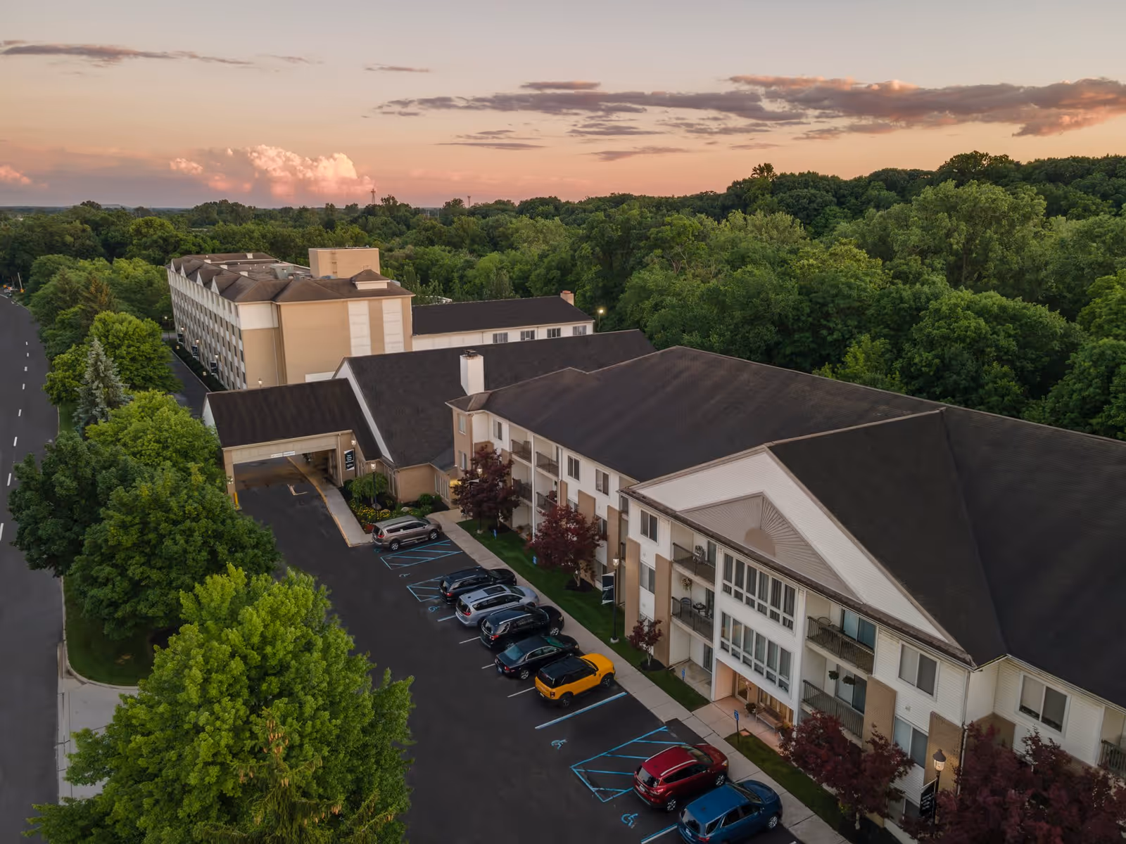 Aerial view of Independence Village of Plymouth senior living facility at sunset, showing a large multi-story building with a parking lot in front and surrounded by lush green trees.