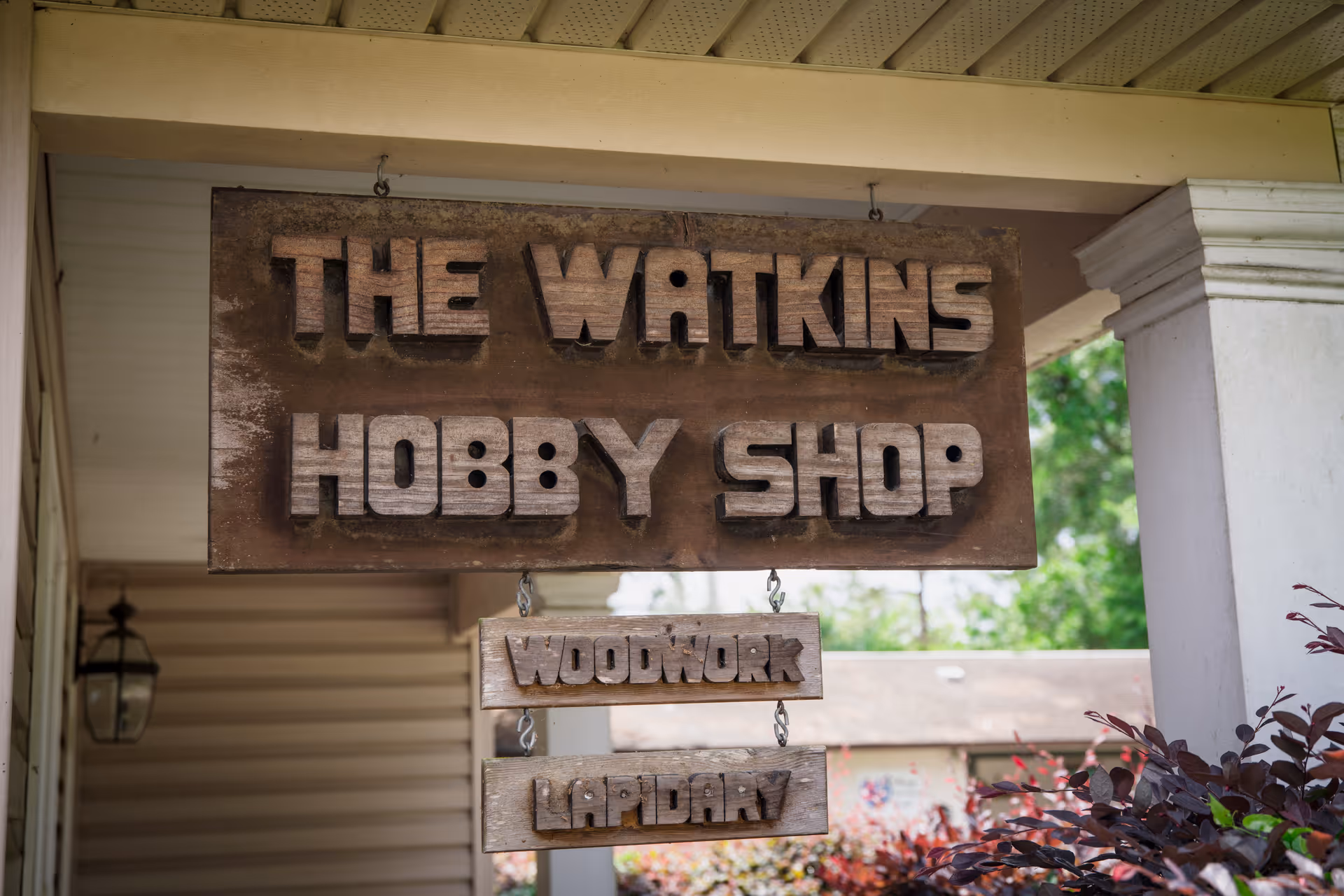 Wooden hanging sign on a porch reading "The Watkins Hobby Shop" with smaller signs below reading "Woodwork" and "Lapidary".