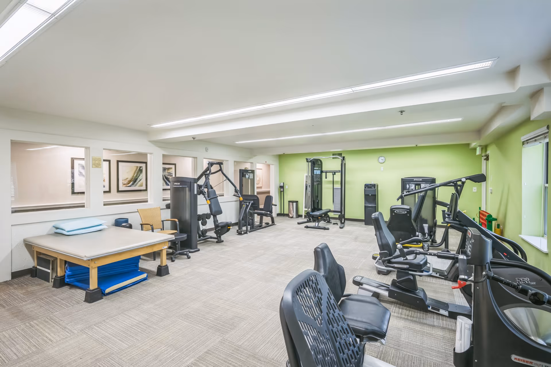 A spacious fitness room with various exercise equipment including weight machines, stationary bikes, and a padded exercise table with pillows. The room has light green walls, large windows on the right side, and bright overhead lighting.