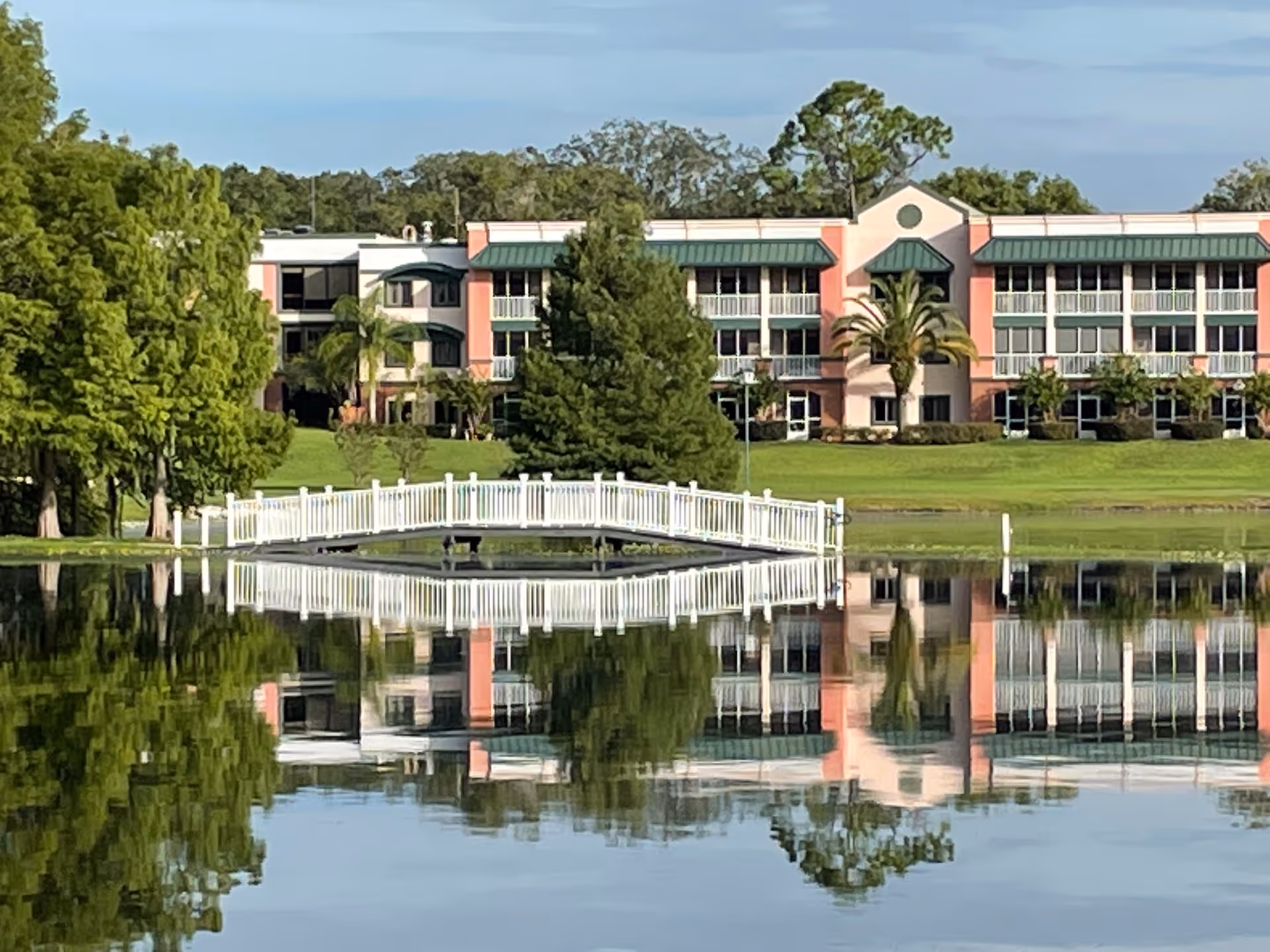 Exterior view of a senior living campus with pink buildings, green landscaping, and a white bridge.