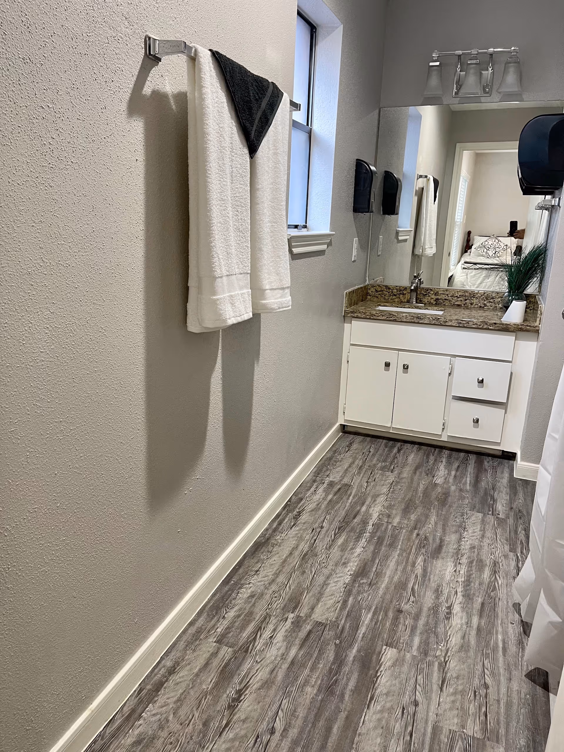 A narrow bathroom with gray wood-look flooring, a white vanity with granite countertop and a sink, a large mirror above the sink, a towel rack on the left wall holding a white towel and a black towel, a small window above the towel rack, and a glimpse of a bedroom visible through the doorway reflected in the mirror.