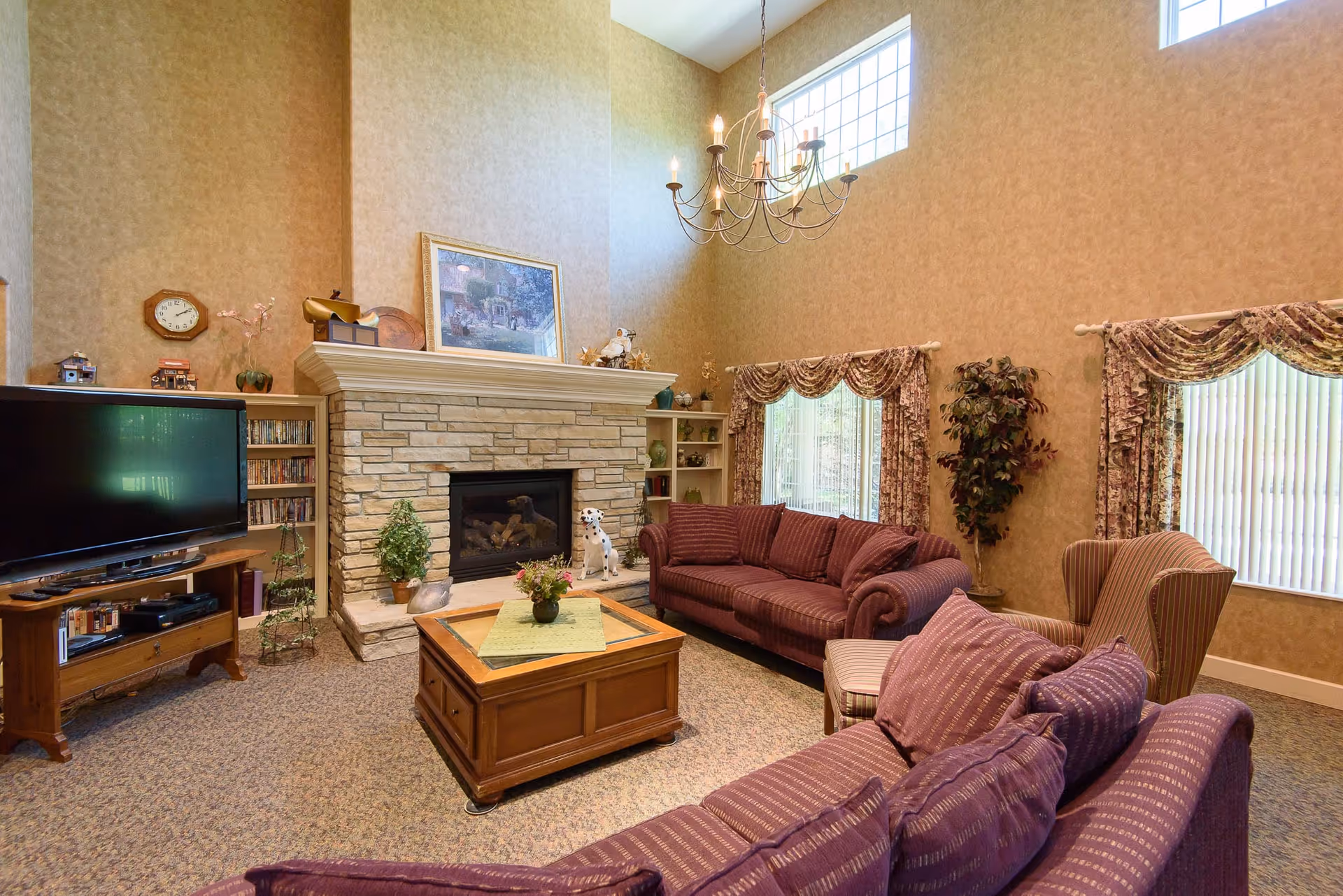 A cozy living room with high ceilings and large windows. The room features two purple sofas, a striped armchair, a wooden coffee table with a small plant centerpiece, a stone fireplace with decorative items on the mantel, and a flat-screen TV on a wooden stand. The walls are beige with patterned curtains on the windows.