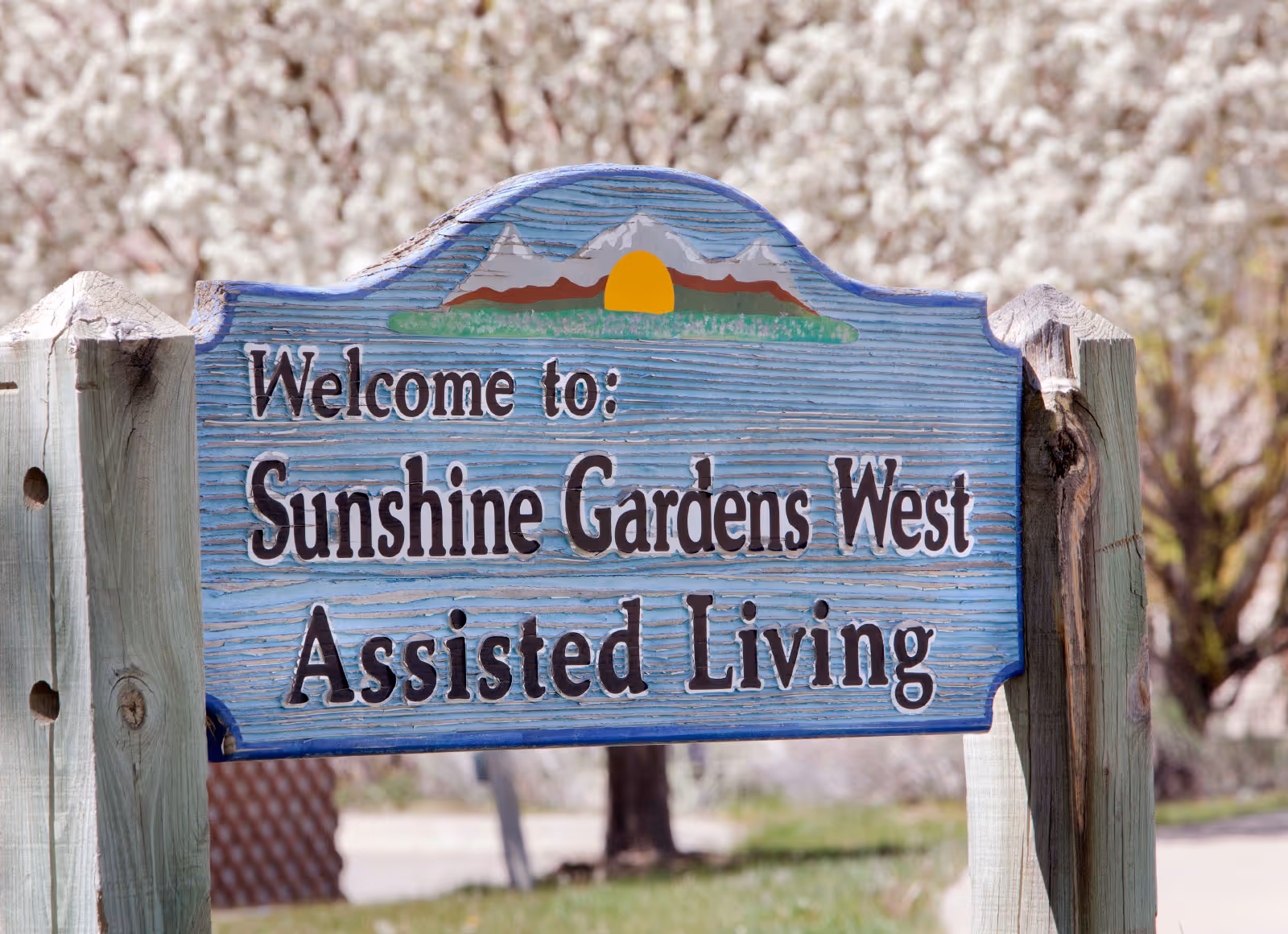 A painted wooden sign reading "Welcome to: Sunshine Gardens West Assisted Living" mounted on posts with flowering trees in the background.