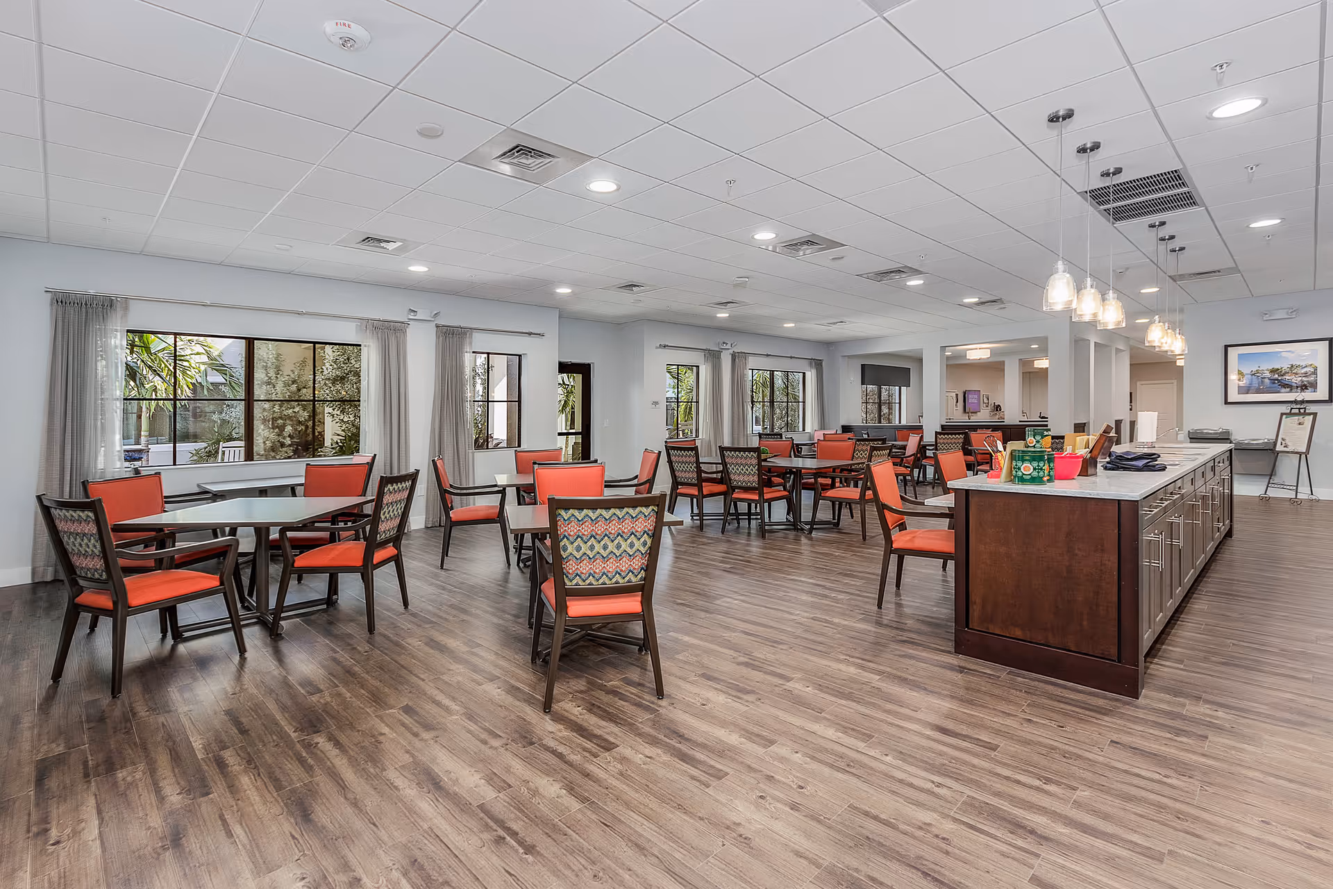 A spacious dining room with multiple tables and chairs featuring red cushions and patterned backs. Large windows with light curtains allow natural light to fill the room. A long island counter with pendant lights hangs above it on the right side, and the floor is covered with wood-style flooring.