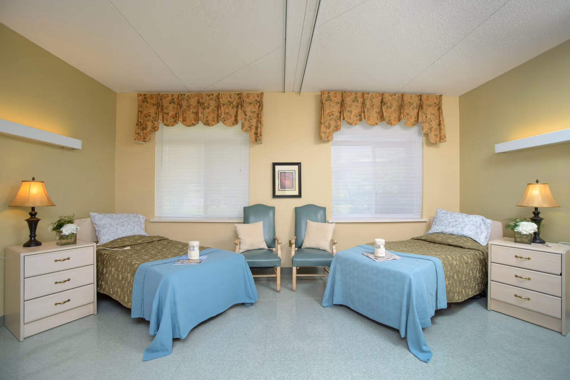 A clean and simple bedroom with two single beds, each with a green bedspread and a blue blanket folded at the foot. Each bed has a white pillow and is flanked by a beige nightstand with a lamp and a small plant. Between the beds are two blue chairs with beige cushions. The room has two windows with floral valances and blinds, and a small framed picture hangs on the wall between the windows.