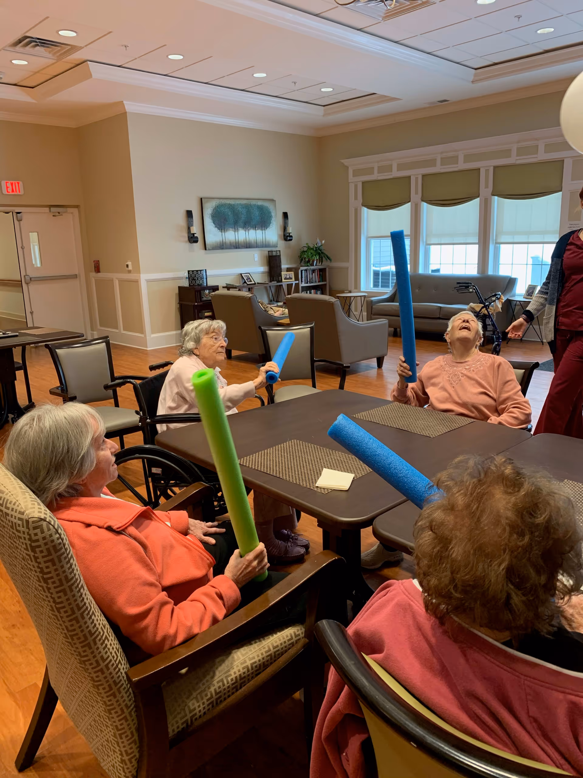 Four elderly women sitting around a table in a well-lit common room, each holding colorful foam pool noodles, engaging in a group activity. The room has comfortable chairs, a couch near large windows with shades, and a painting on the wall. A staff member stands nearby.